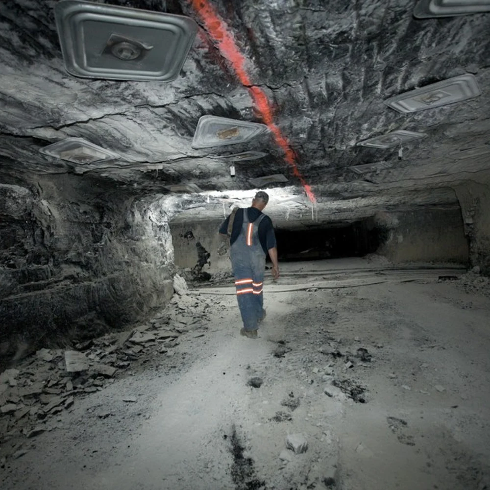 A coal miner in overalls with reflective stripes walking away through a low, rock-walled mine tunnel.