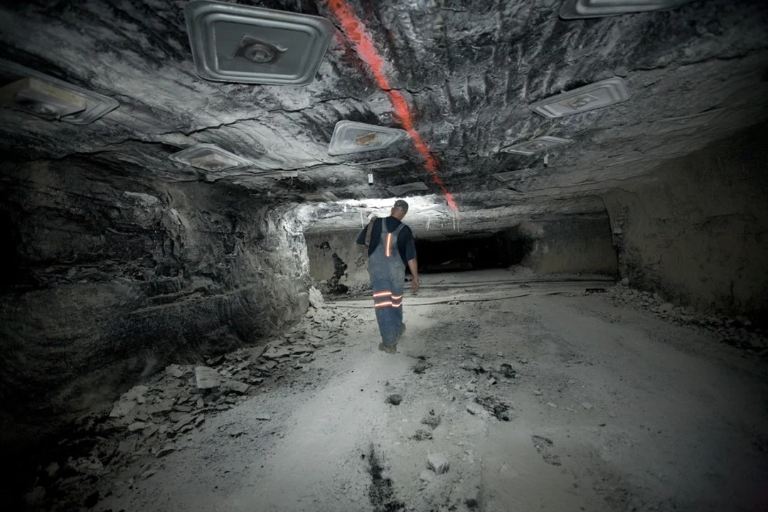 A coal miner in overalls with reflective stripes walking away through a low, rock-walled mine tunnel.