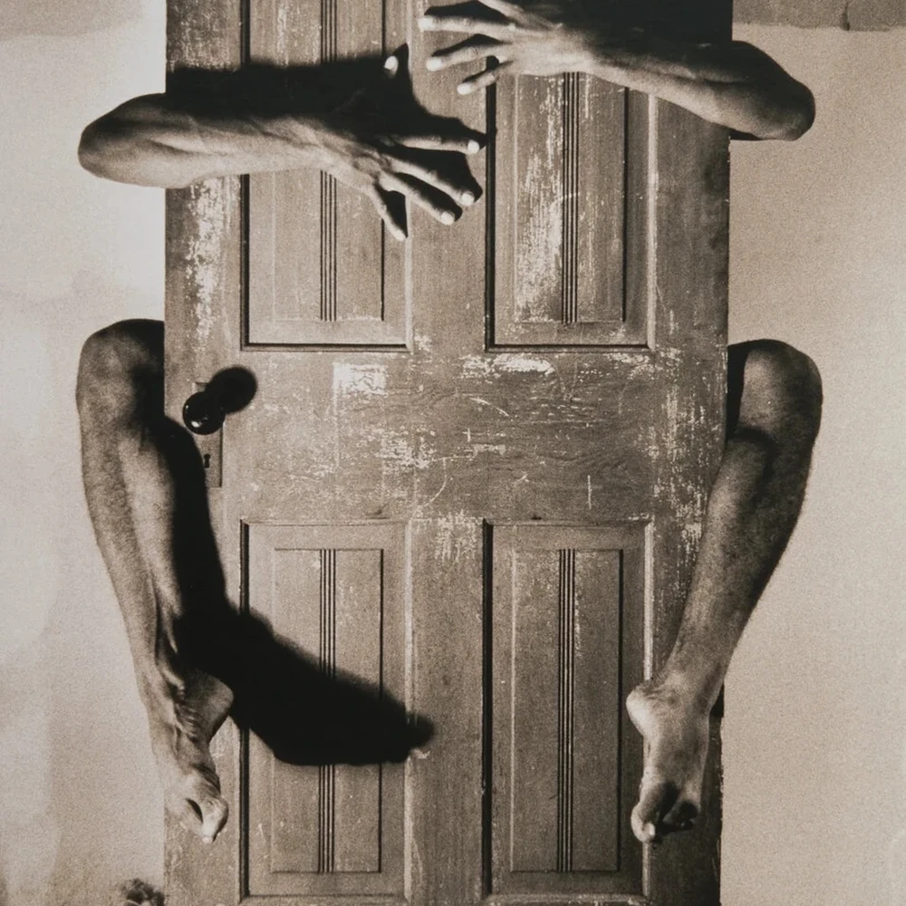 Black-and-white photograph of a wooden door with a pair of arms and legs reaching through gaps around it.
