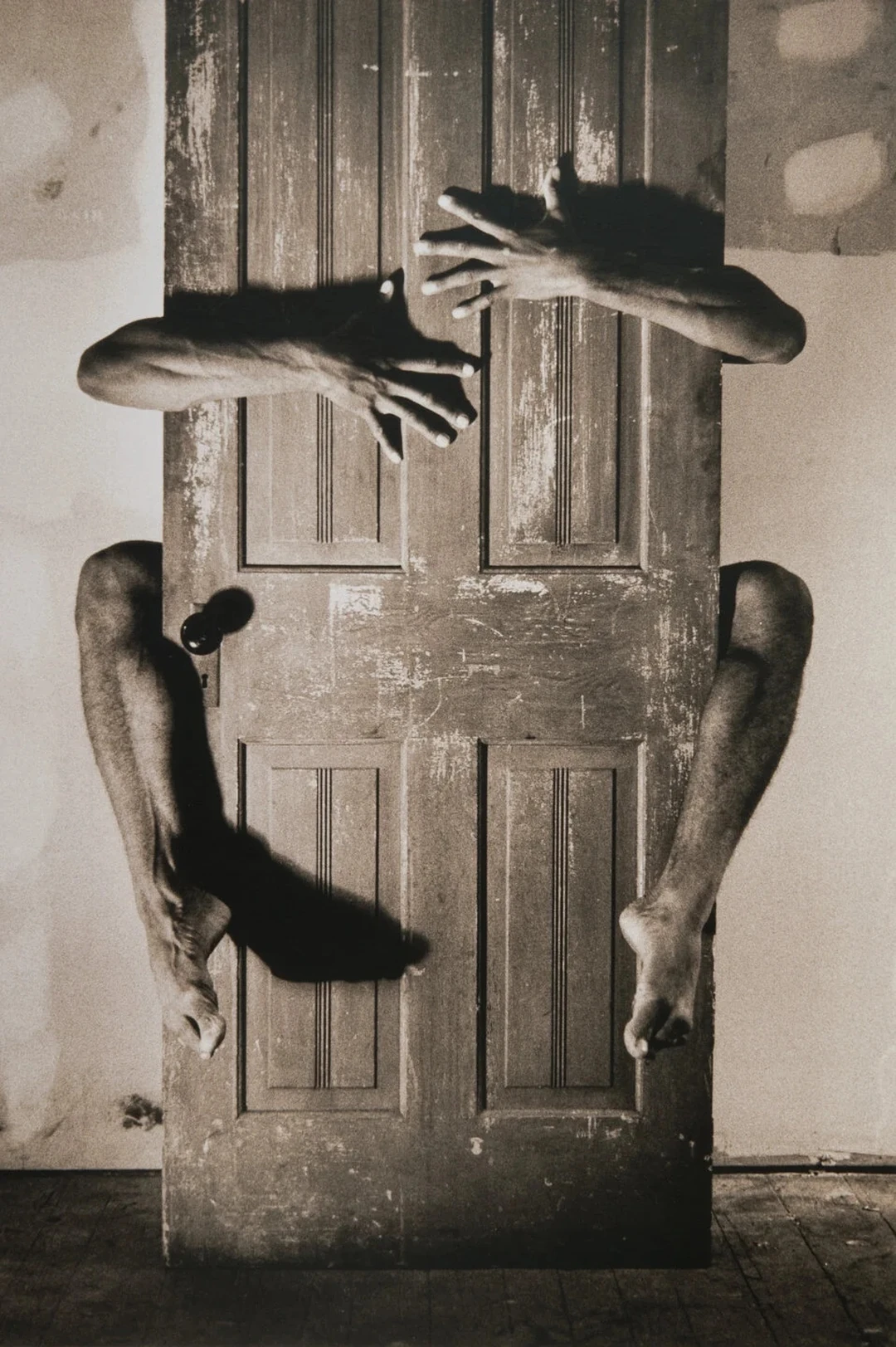 Black-and-white photograph of a wooden door with a pair of arms and legs reaching through gaps around it.
