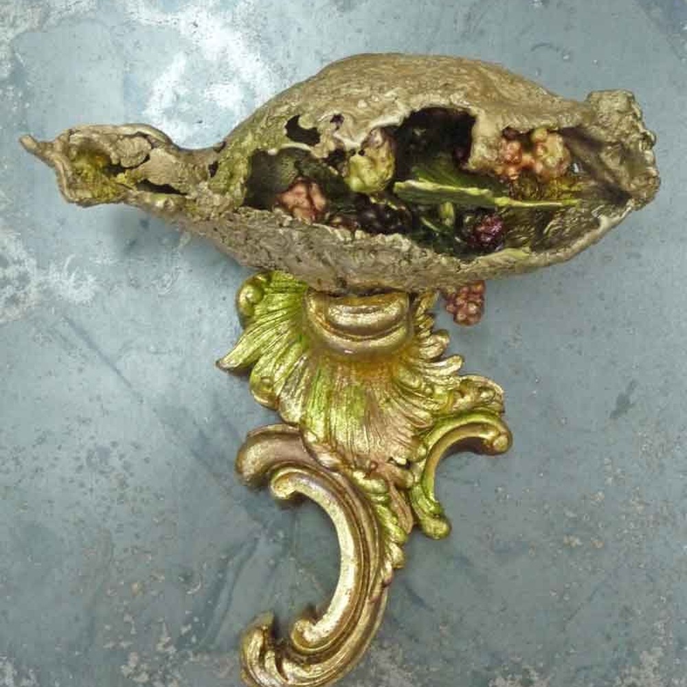 A golden sculptural seed pod, split open to reveal colorful contents, mounted on an ornate gilded scroll bracket against a blue-grey surface.