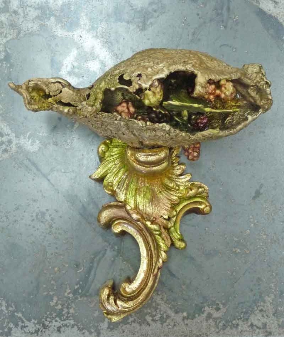 A golden sculptural seed pod, split open to reveal colorful contents, mounted on an ornate gilded scroll bracket against a blue-grey surface.