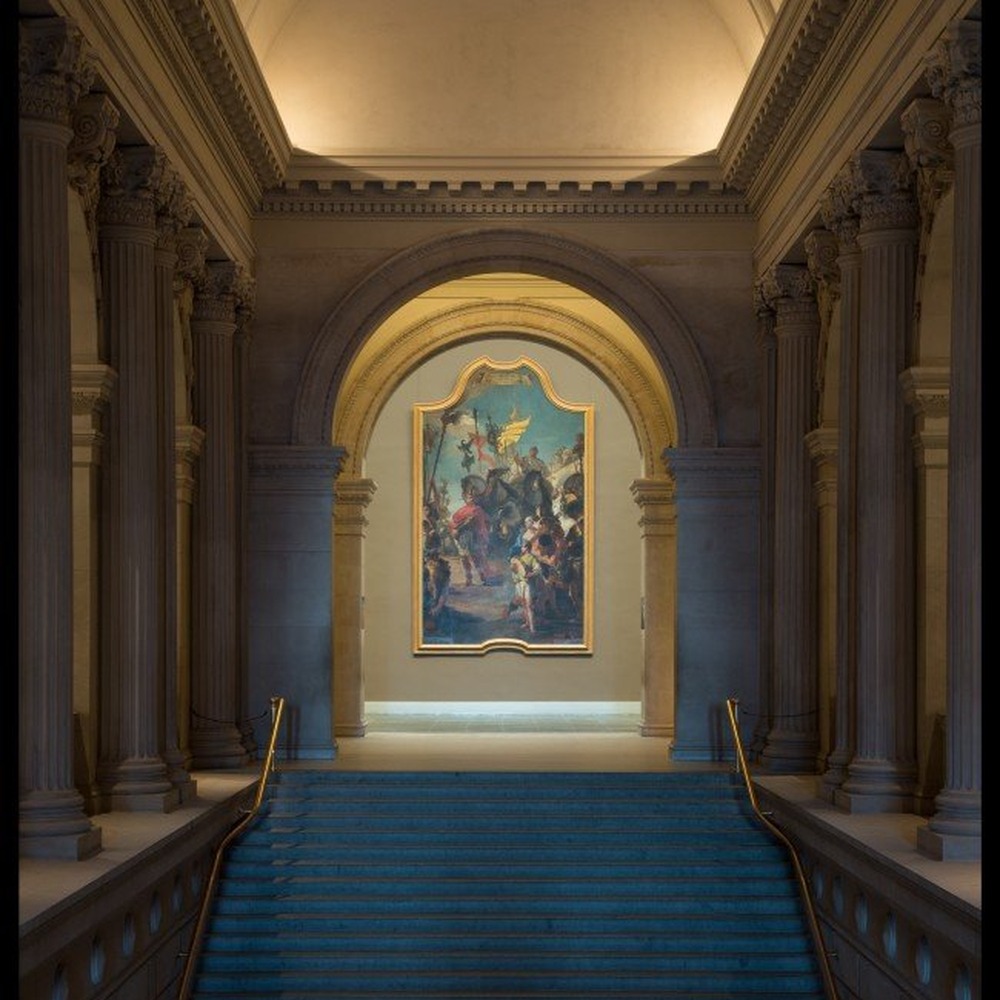 Grand stone staircase with blue carpet leading up to an arched doorway framing a large Tiepolo painting in the Metropolitan Museum's Great Hall.