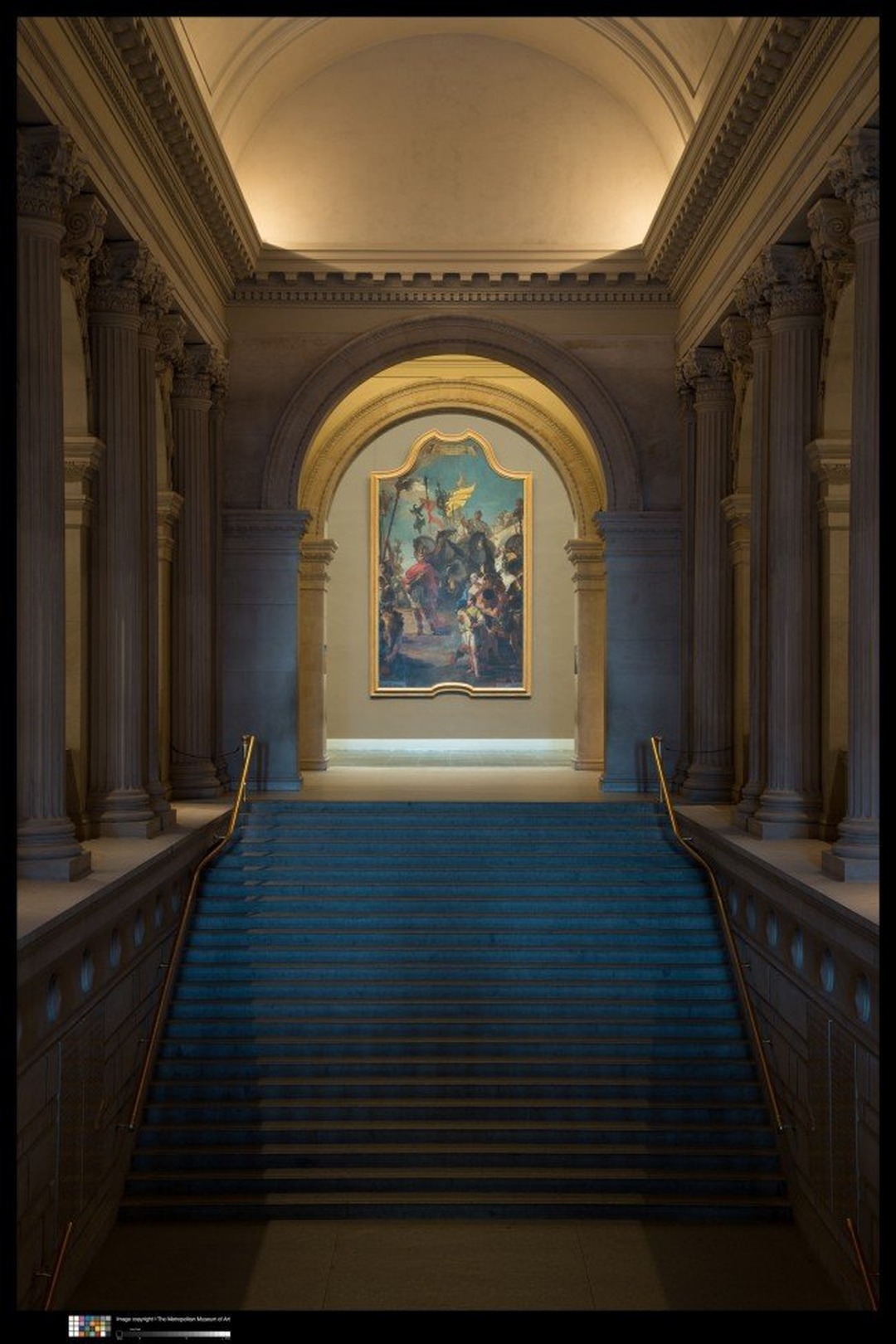 Grand stone staircase with blue carpet leading up to an arched doorway framing a large Tiepolo painting, flanked by tall columns in the Metropolitan Museum's Great Hall.