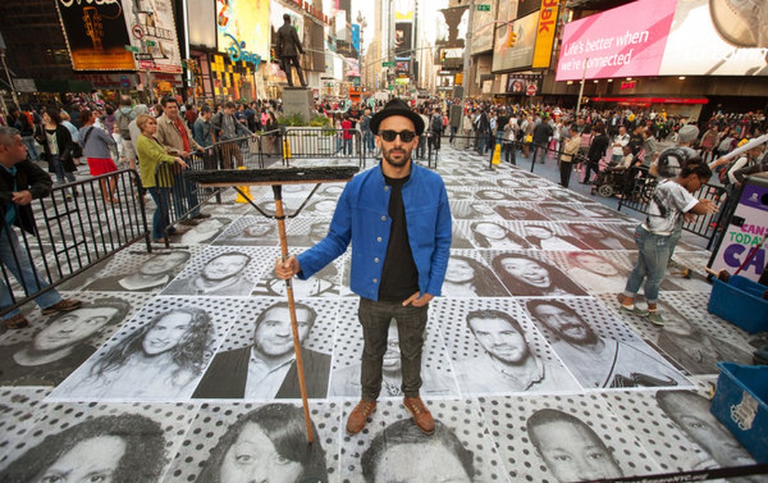 The artist JR, wearing a blue jacket, black shirt and a hat, holding a long-handled squeegee, standing in the middle of a pedestrian plaza in Times Square covered with large black-and-white paste-up portraits, part of his Inside Out project.