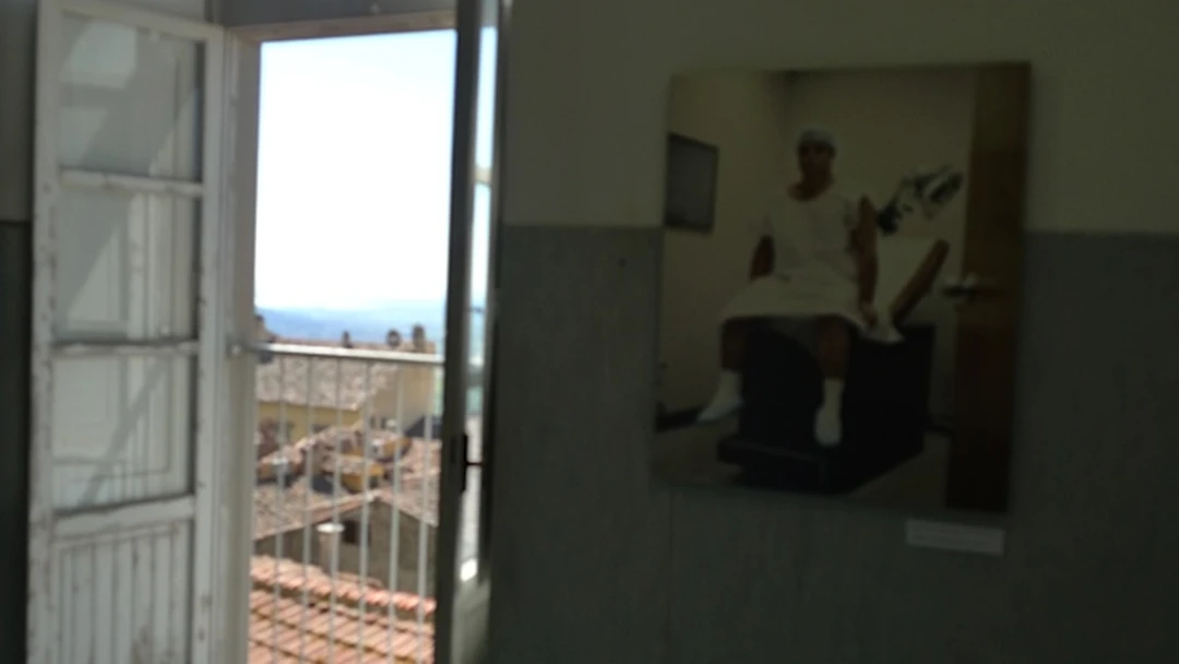 Installation view inside the ancient hospital in Cortona: a framed photograph of a seated figure on a wall beside an open doorway with a view over tiled rooftops.