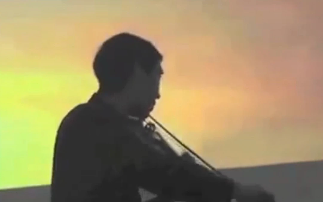A violinist silhouetted in dark profile, playing against a glowing yellow-orange projected backdrop.