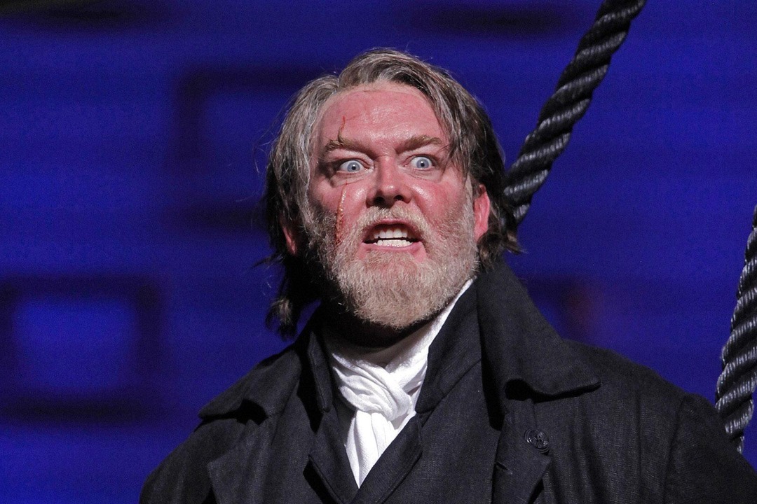 Jay Hunter Morris as Captain Ahab, close-up on stage in a dark captain’s coat with ship’s rigging beside him, against a deep blue backdrop.
