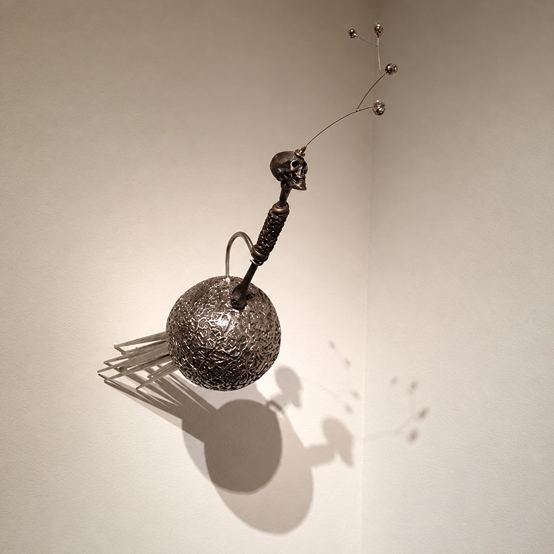 Round, textured metal sphere with a small skull on a stalk, a curved wire and two small balls, mounted in the corner of a white wall and casting soft shadows.