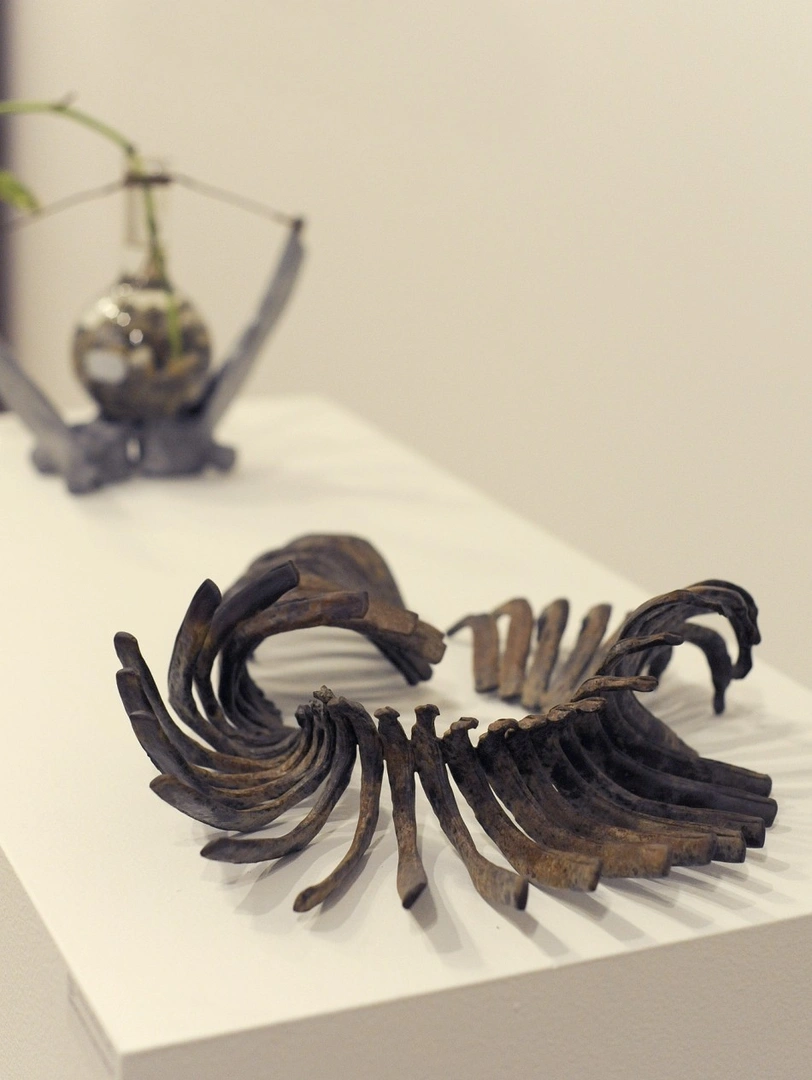 Curved sculpture of rib-like dark metal forms on a white pedestal, with a small hanging glass-sphere sculpture behind it.