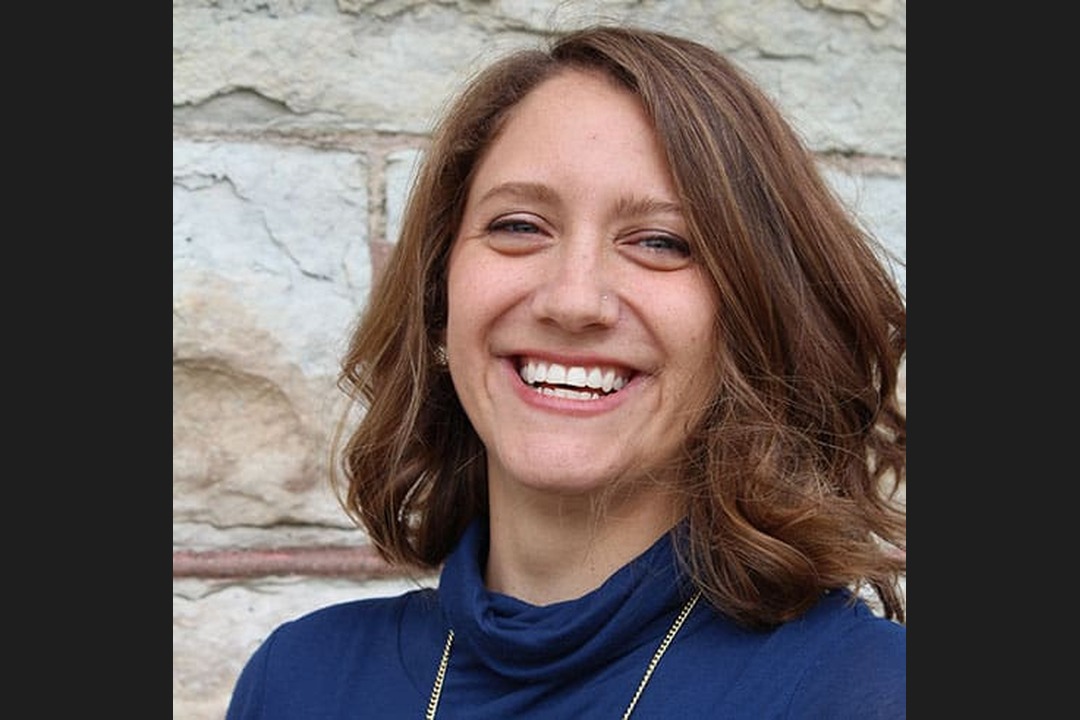 Headshot of Leslie Mooney, a smiling woman with shoulder-length brown hair wearing a blue top and a gold necklace, against a stone wall.