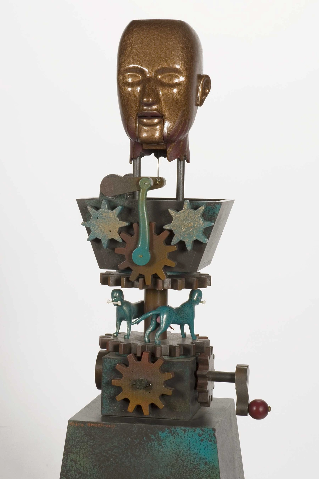 Dogs of War detail 42 x 13 x 13. Steve Armstrong: gilded head of a male figure above a mechanism of gears and small dogs on a green pedestal.