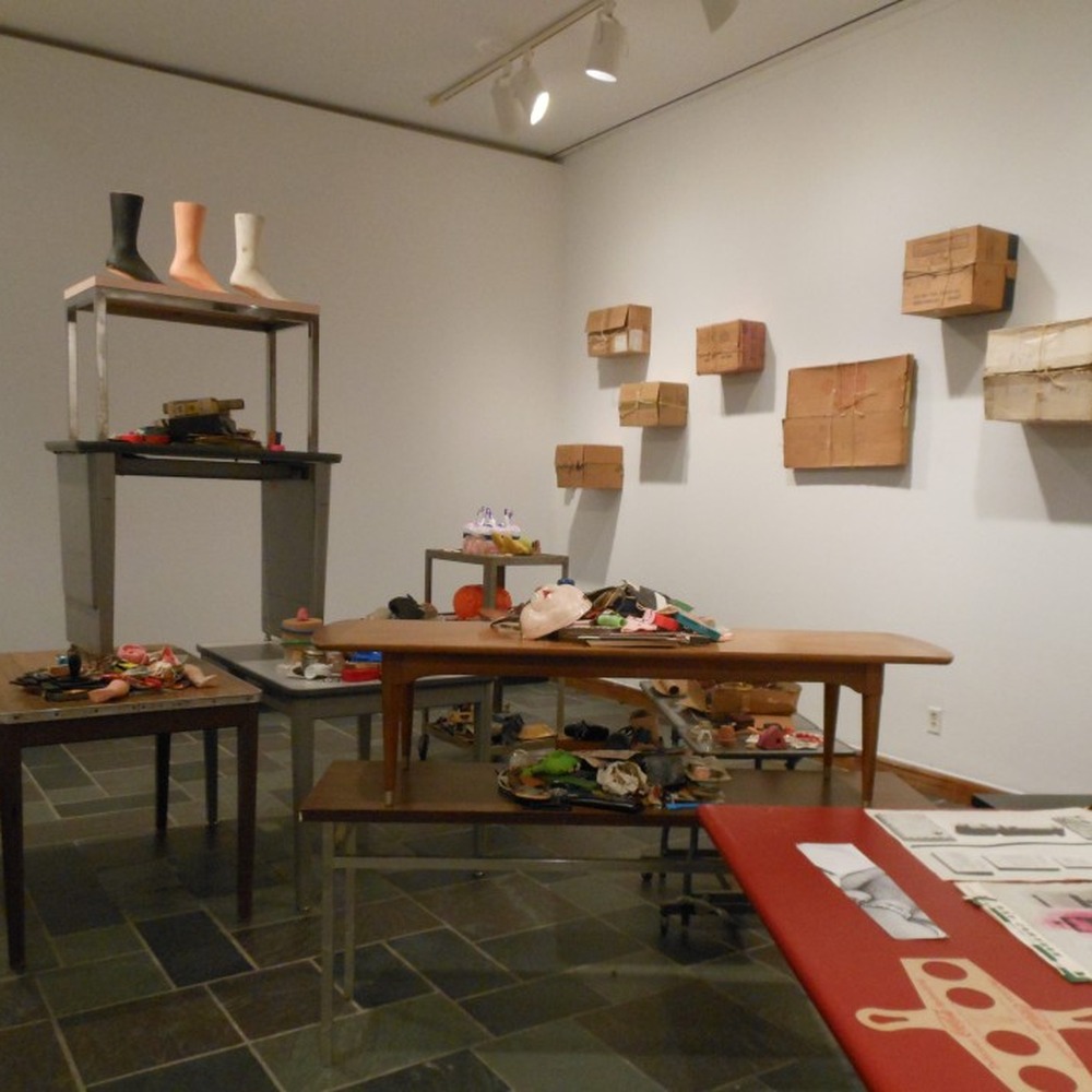 Installation view of the Bob Boxes exhibition: tables covered with small objects and wrapped packages, with cardboard boxes tied with string mounted on the gallery walls.