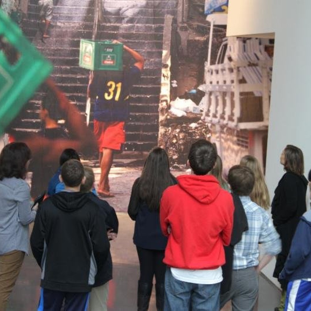 A group of visitors, many of them young people, stands in a gallery looking at a wall-size photograph of a man in a number 31 shirt and red shorts carrying a green crate on his head up a flight of outdoor stairs.