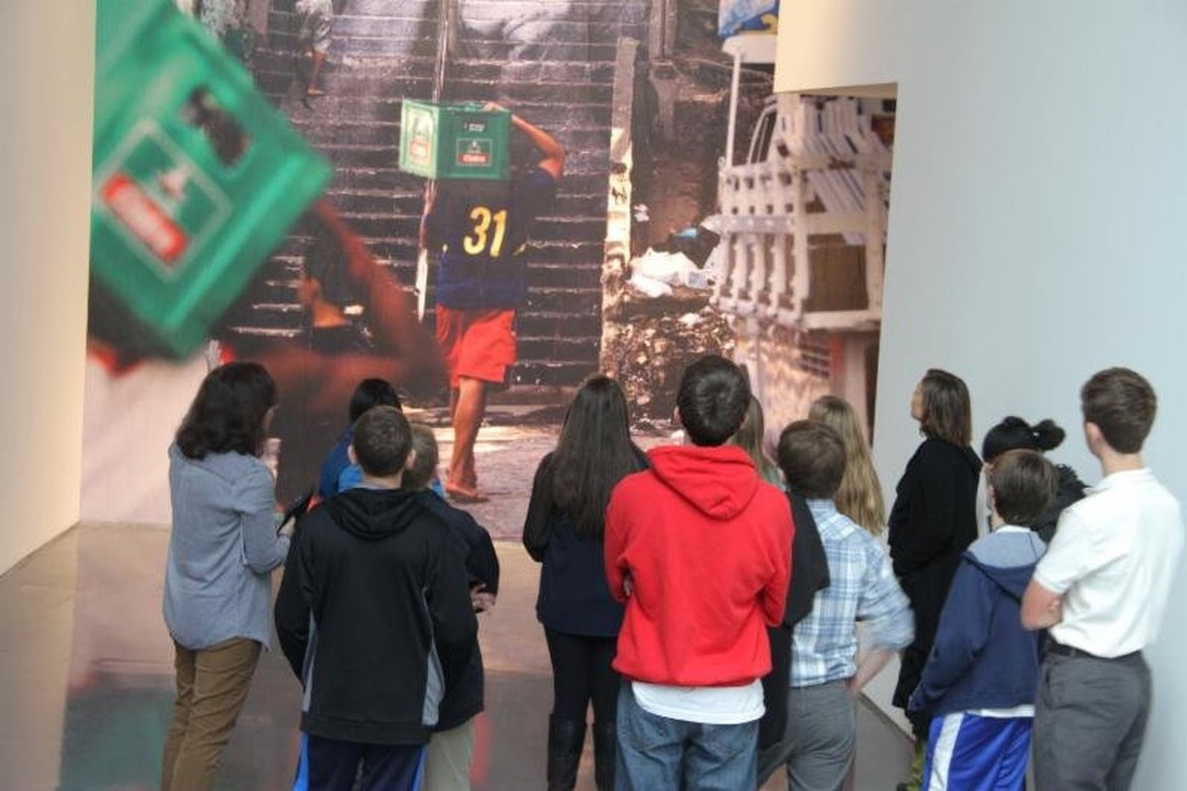 A group of visitors, many of them young people, stands in a gallery looking at a wall-size photograph of a man in a number 31 shirt and red shorts carrying a green crate on his head up a flight of outdoor stairs.