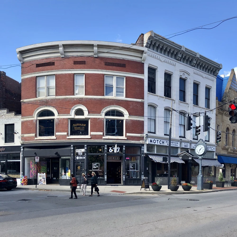 Downtown Covington, Kentucky, at the corner of Pike Street and Madison Avenue: historic brick and stone commercial buildings with shops at street level, a street clock on the sidewalk and pedestrians crossing the street on a clear day.