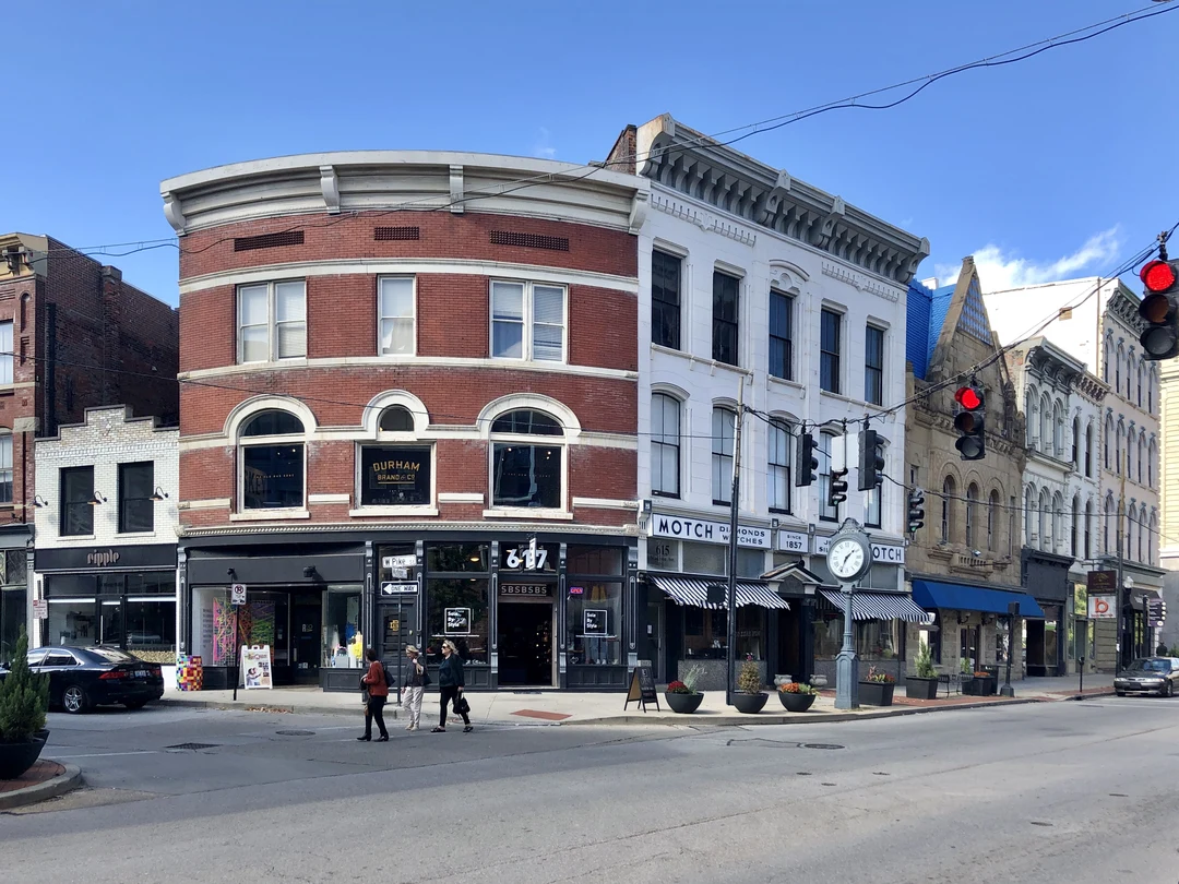 Downtown Covington, Kentucky, at the corner of Pike Street and Madison Avenue: historic brick and stone commercial buildings with shops at street level, a street clock on the sidewalk and pedestrians crossing the street on a clear day.