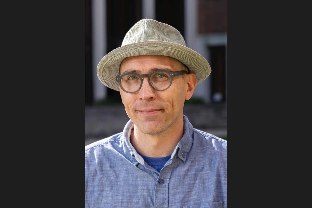 Portrait of artist Michael Stillion wearing a straw hat and glasses, outdoors.