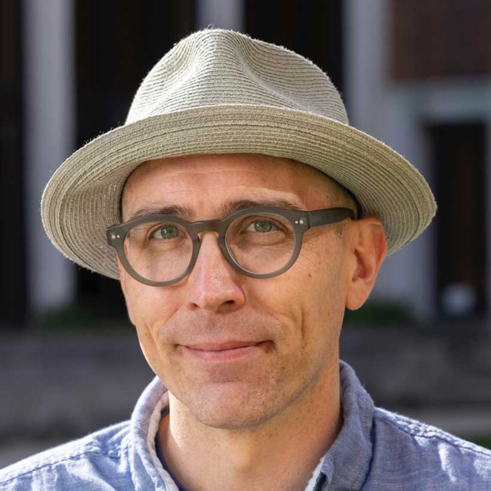 Portrait of artist Michael Stillion wearing a straw hat and glasses, outdoors.