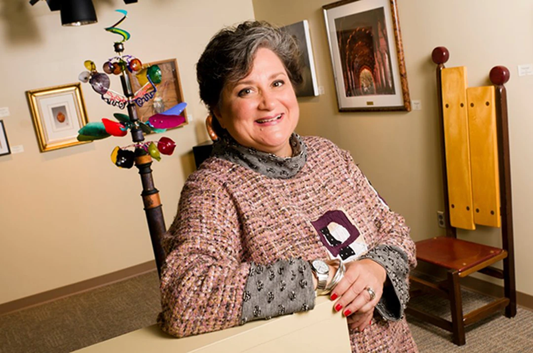 Portrait of Sharon Strubbe, smiling, in an interior with framed artworks, a colorful hanging sculpture and a wooden chair.