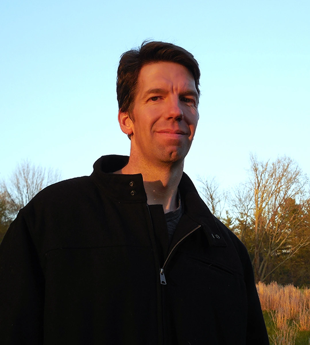 Portrait of Kevin Muente, smiling, outdoors at sunset in a dark jacket, with bare trees behind him.