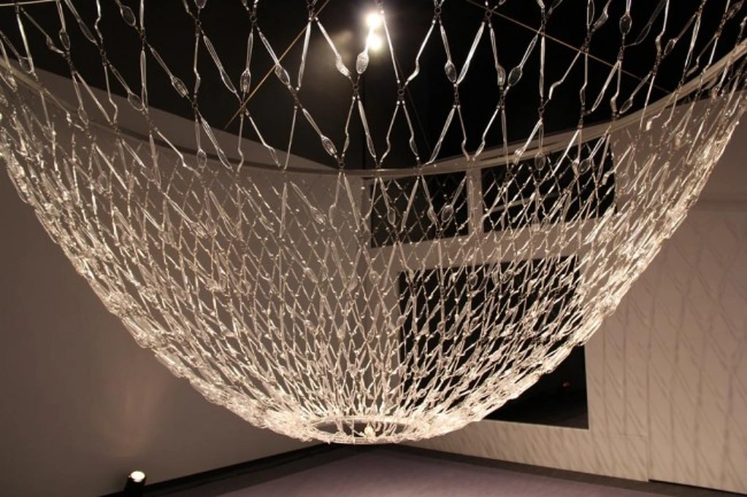 Suspended sculpture of interlaced clear plastic pieces, shaped like a bowl and seen from below in a gallery.