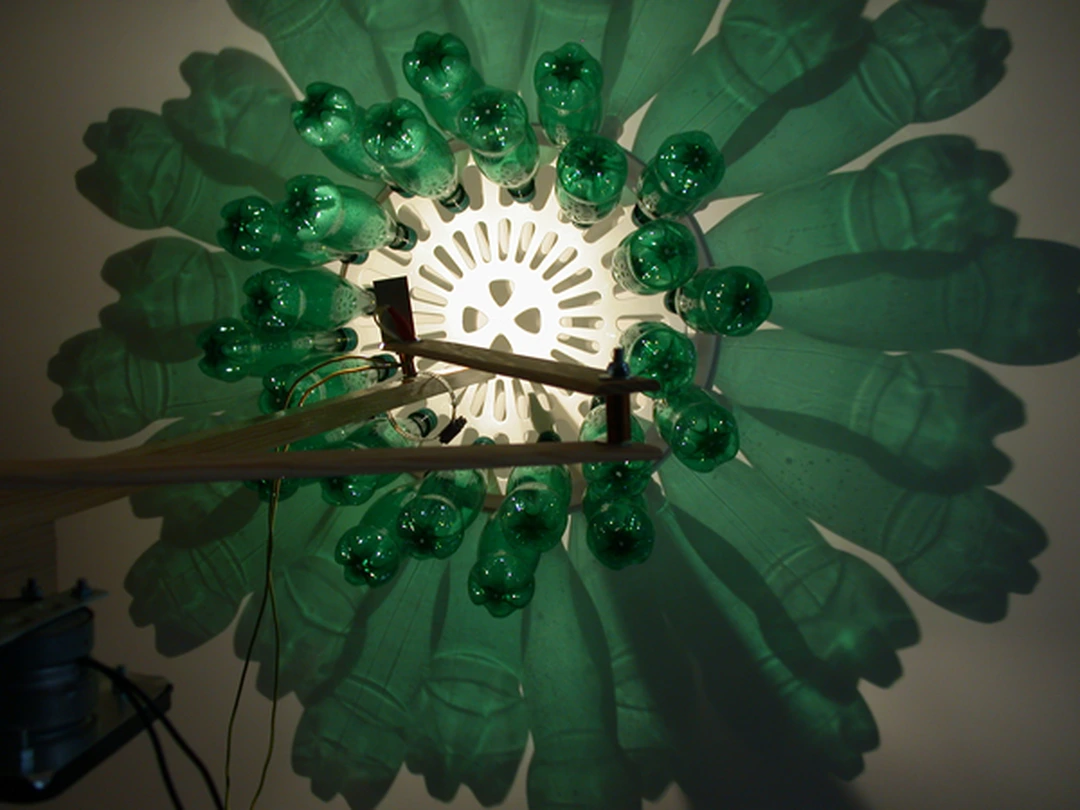 Circular arrangement of green plastic bottles around a central light, casting shadows on the wall.