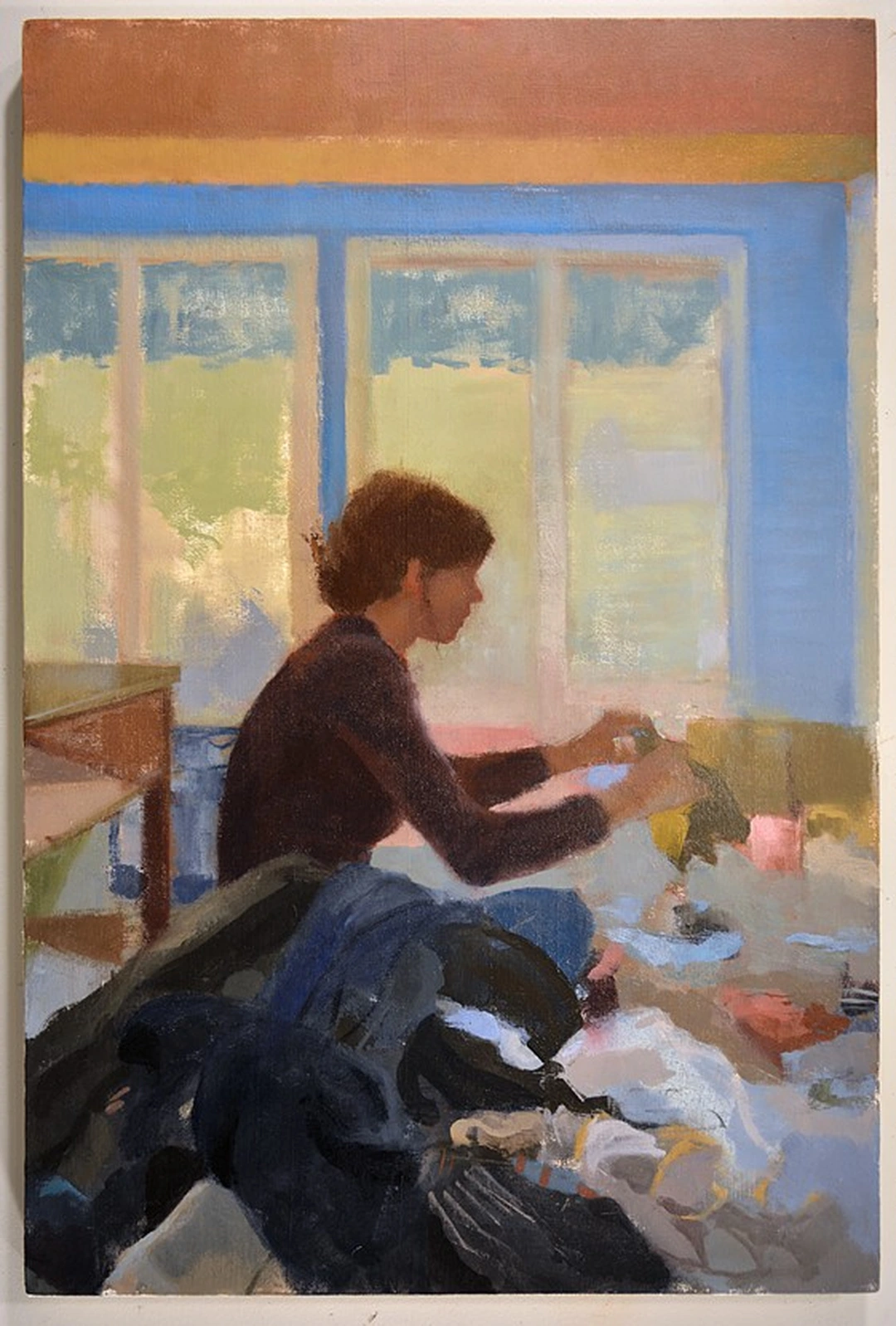 Oil painting of a woman in a maroon sweater sitting by a bright window and sorting a pile of laundry, with blue window frames and a dark jacket in the foreground.