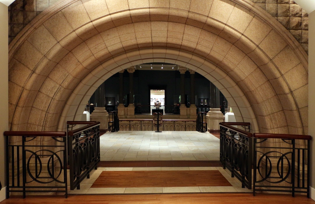 Stone archway with a curved coffered ceiling frames a stair landing with metal railings, and a gallery with columns visible beyond.