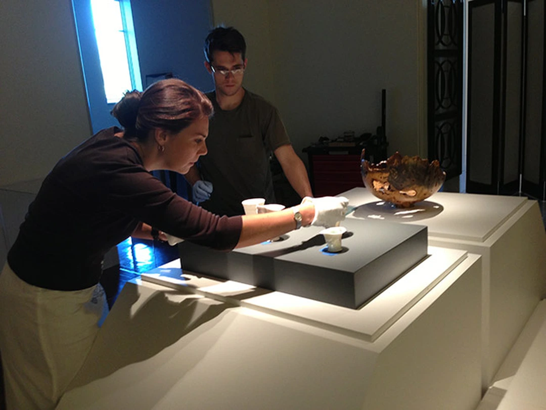A woman in white gloves places a small cup in a display case beside a bronze bowl, while a man stands behind her in a dim gallery.