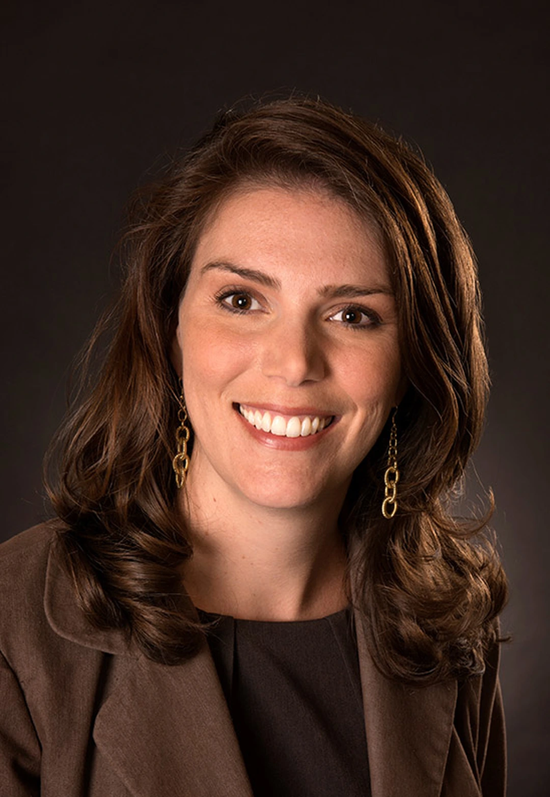 Portrait of a smiling woman with long brown hair and a brown jacket, against a dark background.