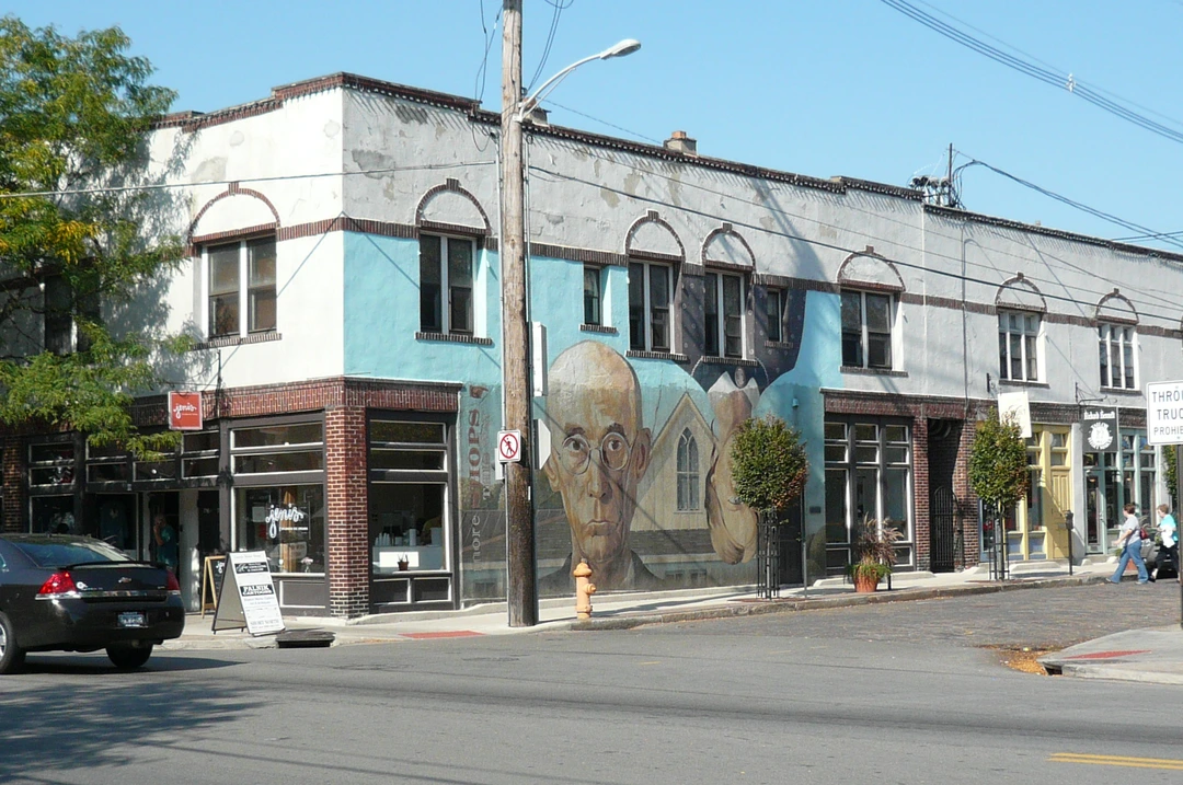 Painted mural of a man in round glasses on the side of a light blue and white brick building, with shops and a sidewalk below.