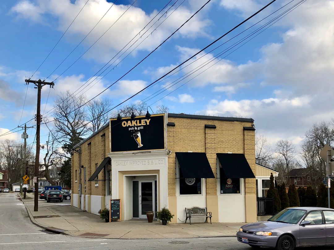 Yellow-brick corner building with a black Oakley sign above dark awnings, on a sidewalk in Oakley with a car parked nearby.