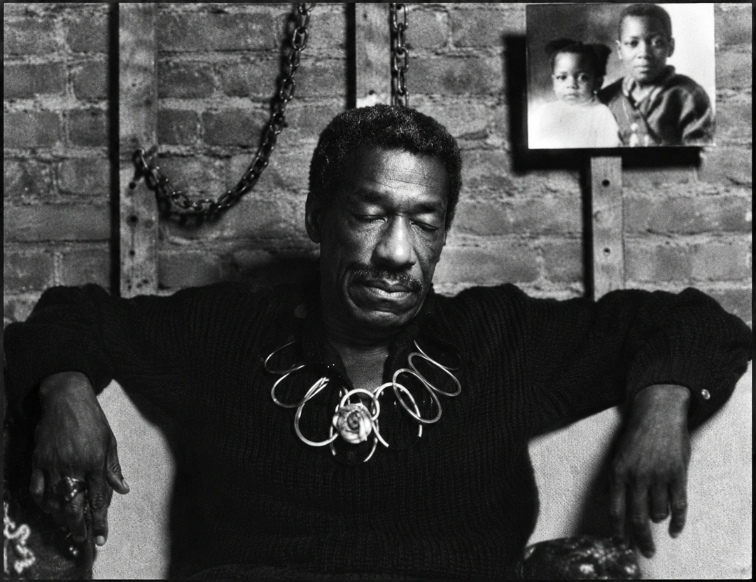 Black-and-white photograph of a seated man in a dark sweater and wire necklace, in front of a brick wall with framed photographs.