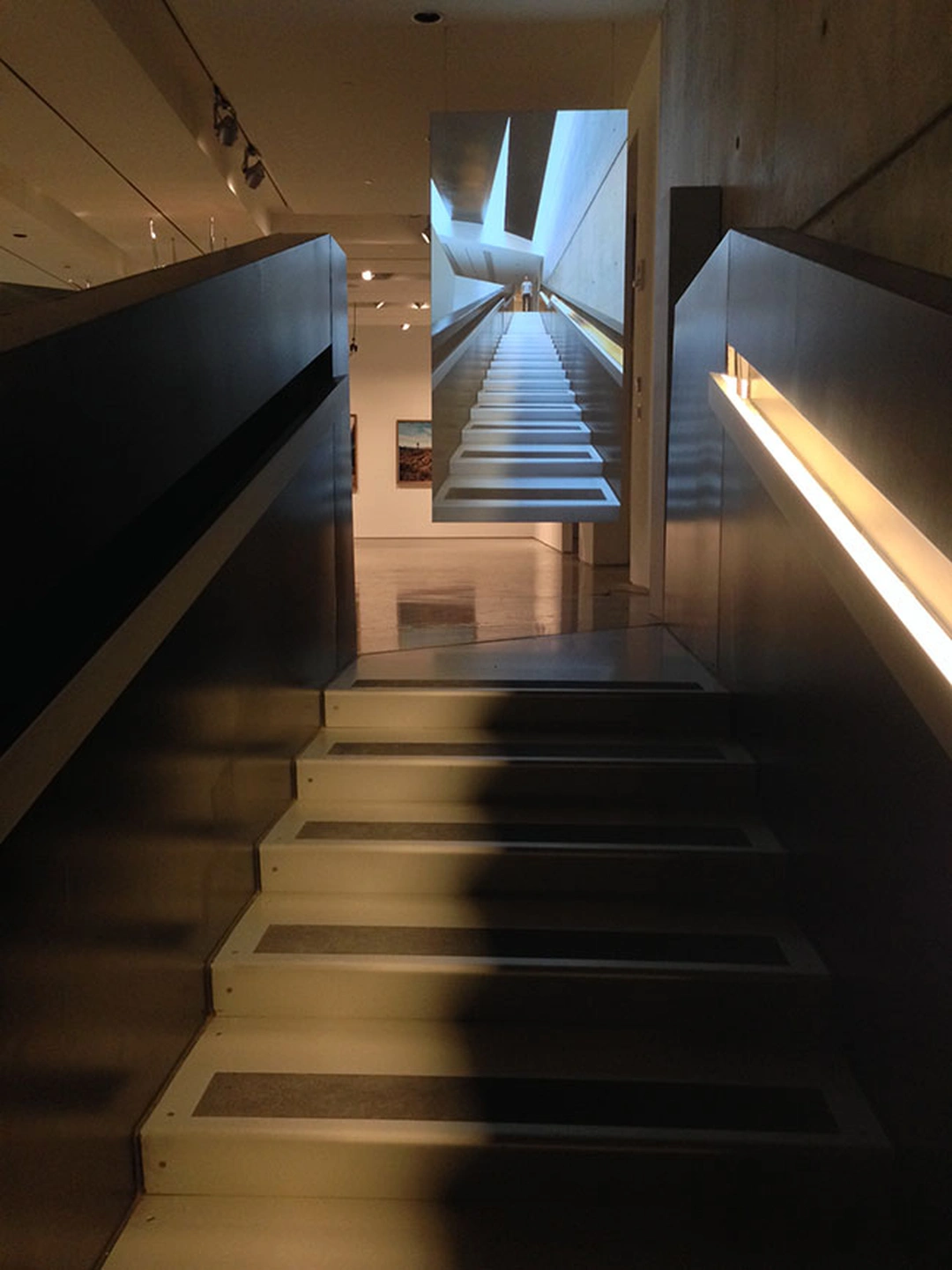 A dark staircase with lit metal treads leading up toward a reflective panel, with a hallway and a framed photograph visible at the top.