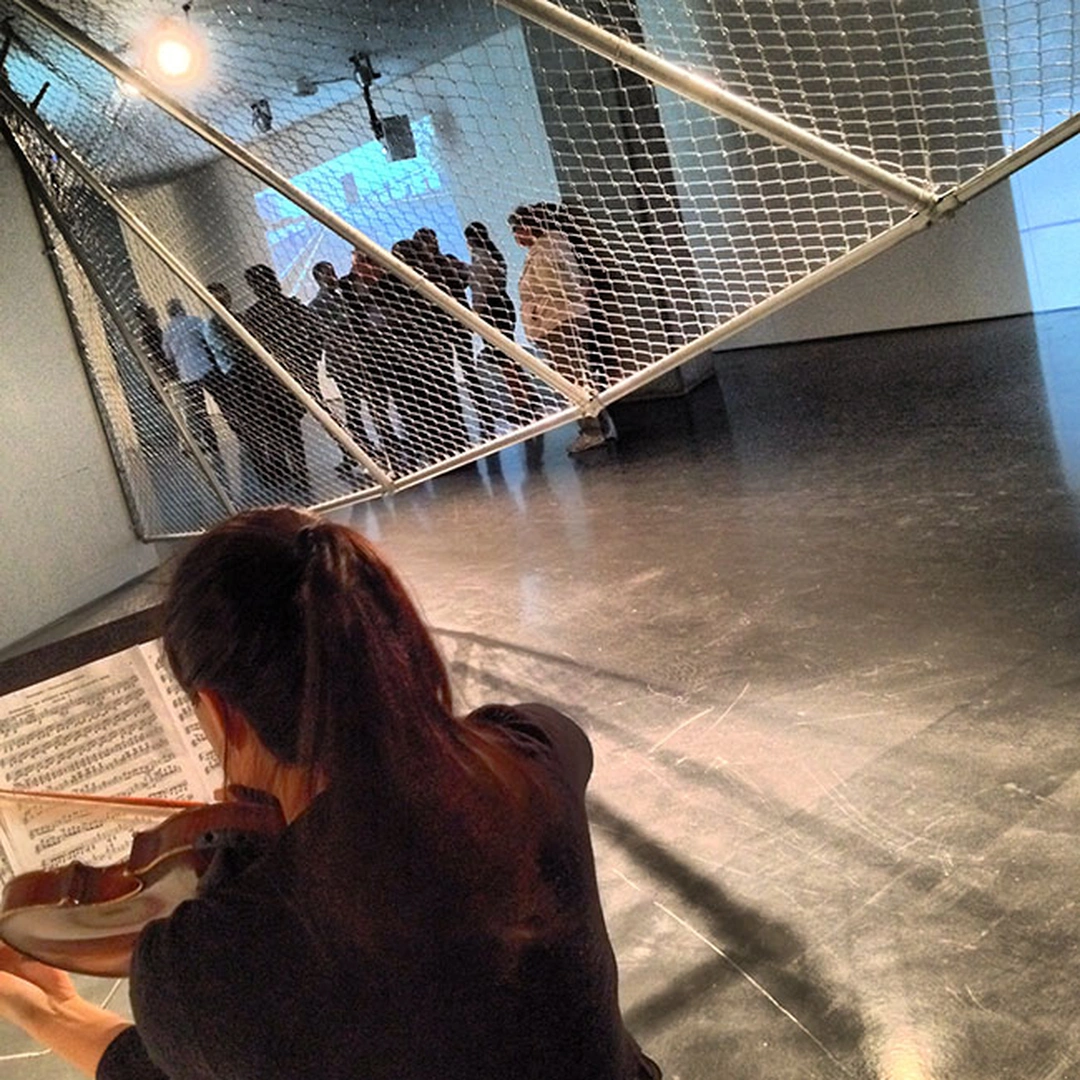 A violinist seen from behind in the foreground, reading sheet music, with a metal mesh overhead and people standing behind it.