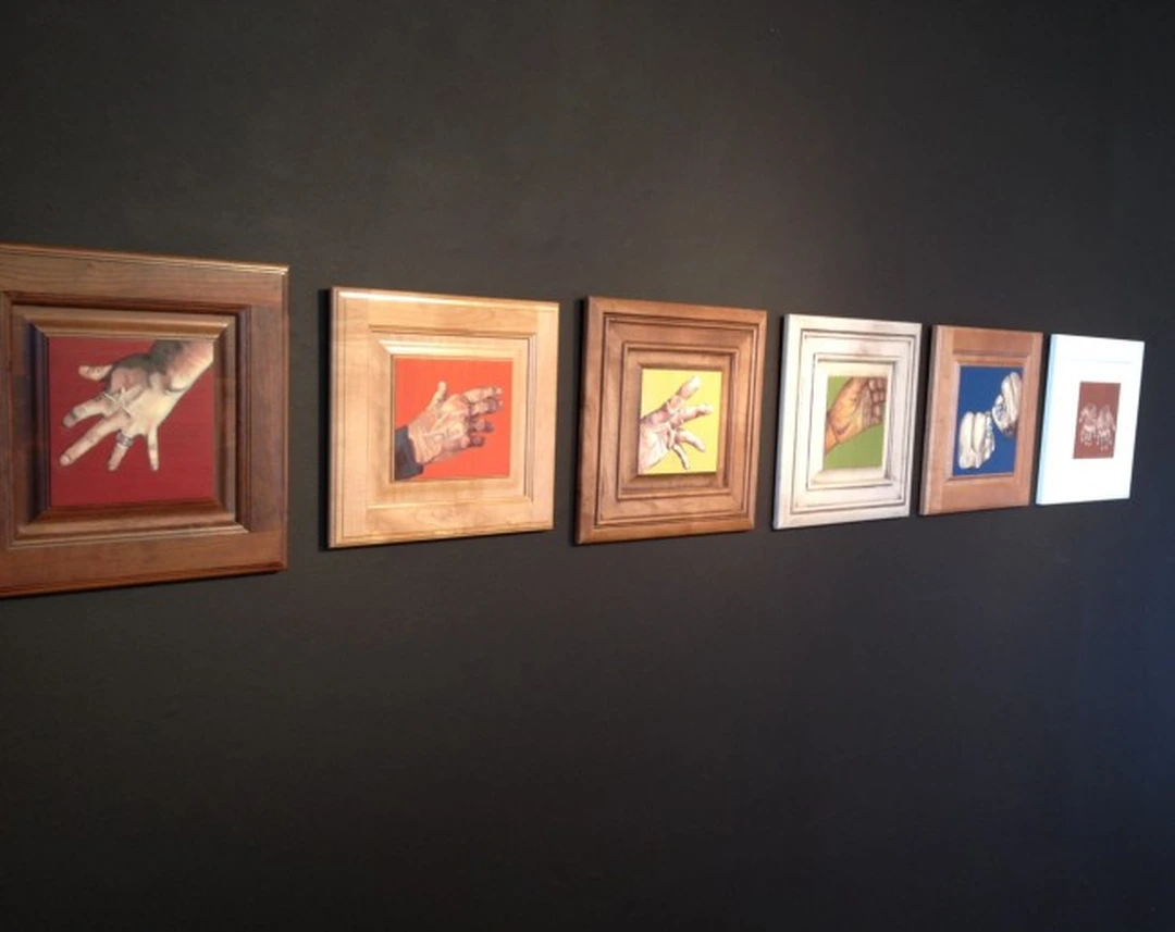 Six small framed paintings of hands in a row on a dark gallery wall.