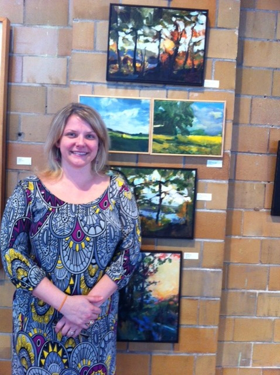 Smiling woman with her hands clasped, standing in front of framed landscape paintings on a brick wall.