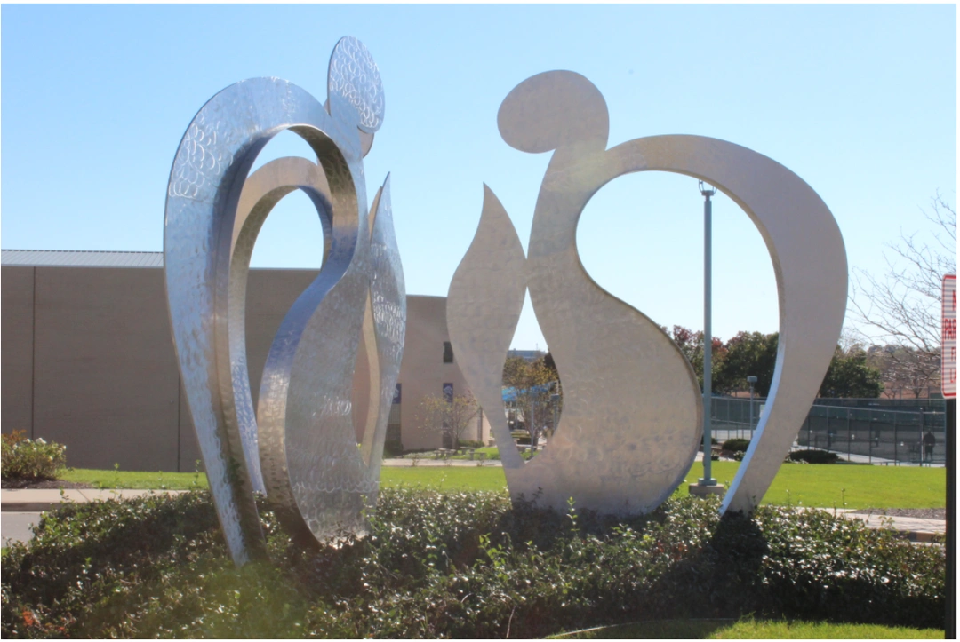 Two abstract stainless steel figures on a shrub bed, with a low building and a lawn behind them under a clear sky.