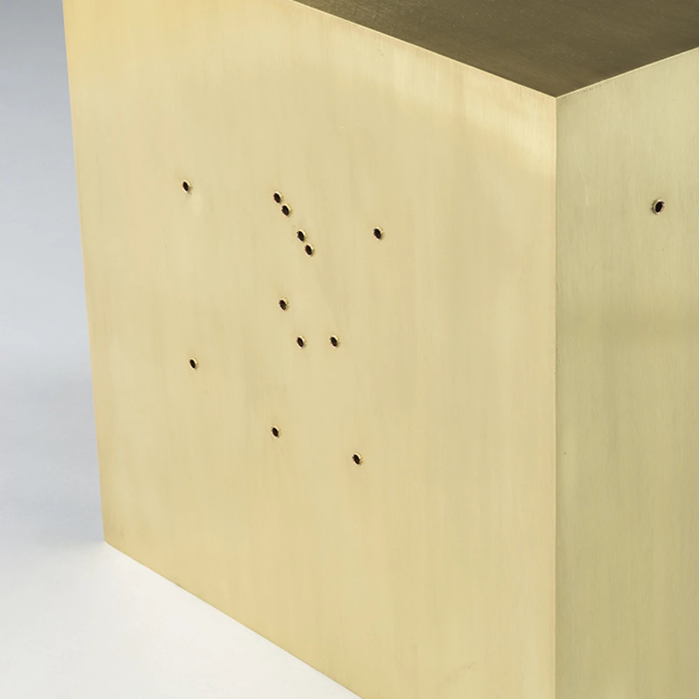 Close-up of a brass cube with a cluster of bullet holes across its front face.