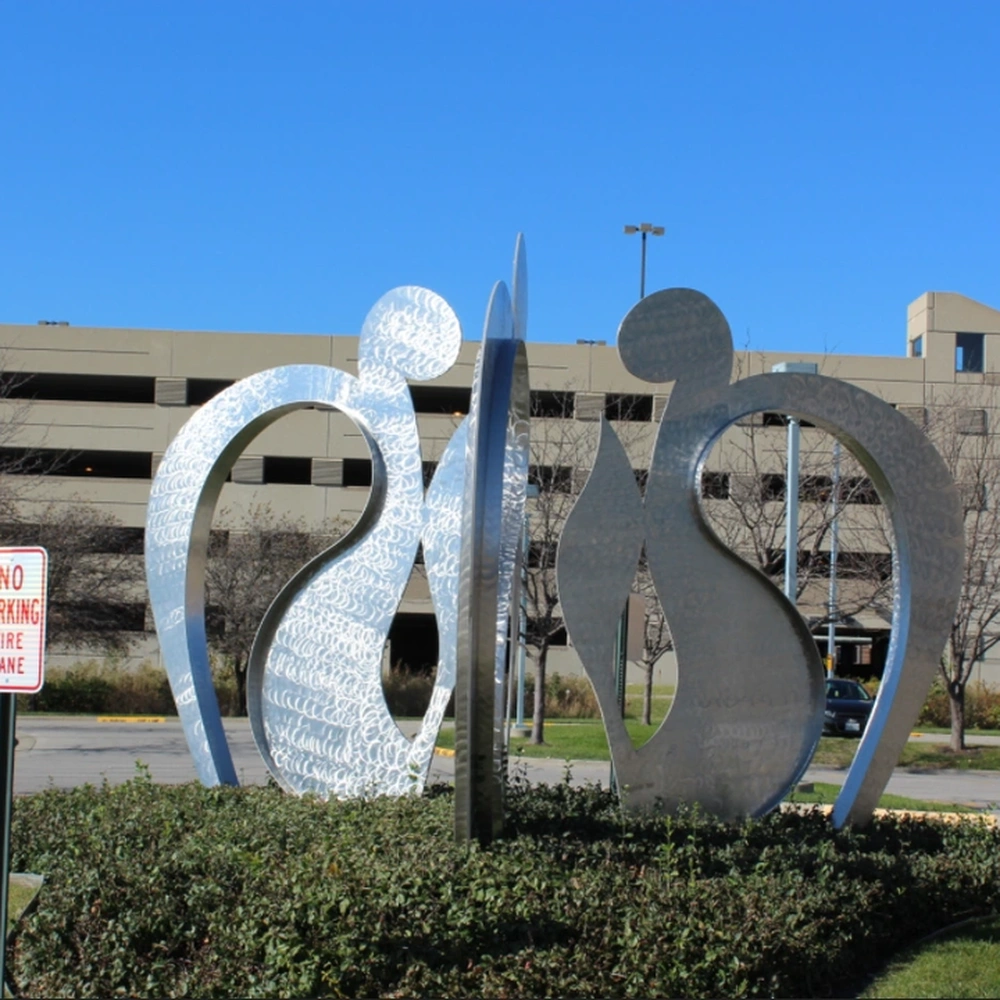 Two abstract stainless steel figures on a shrub bed, one with a hammered, scalloped surface, with a parking structure and a clear blue sky behind them.