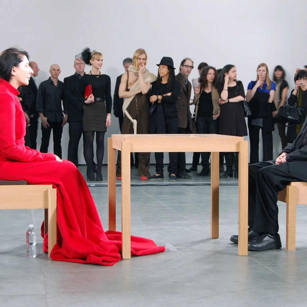 Photograph of Marina Abramović in a red dress seated across a light wooden table from a man in black.