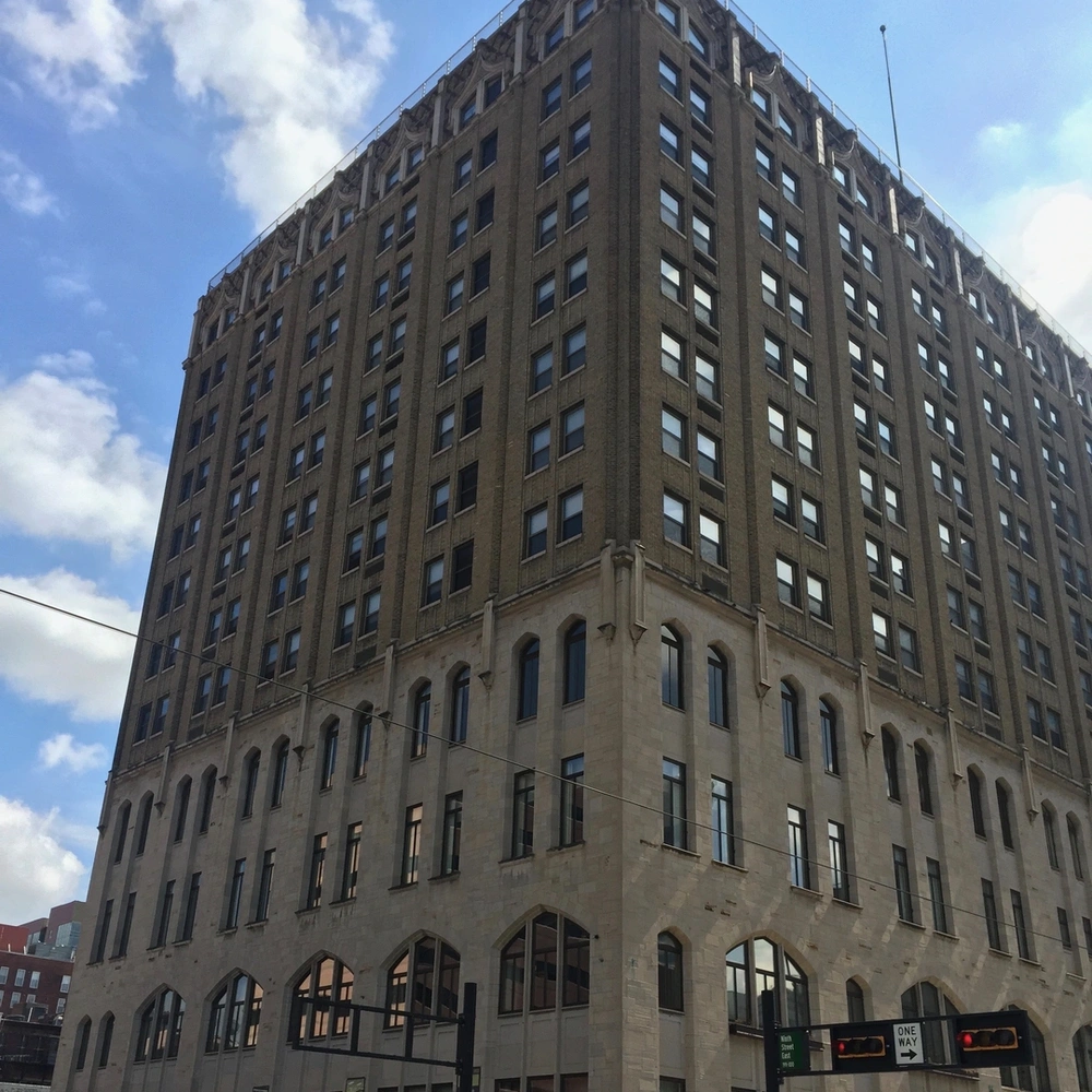 Photograph of a tall beige brick office building with rows of windows and arched ground-floor openings on a city street under a blue sky.