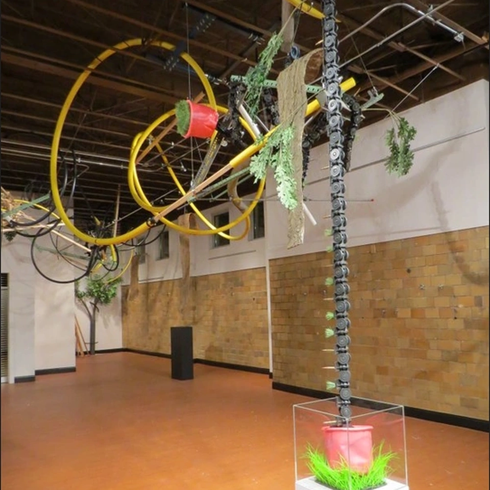 Installation of yellow plastic tubes, hoops and hanging plants filling the upper space of a gallery with a wooden floor, with an artificial tree rising from a clear box with grass and a red pot on a white pedestal.
