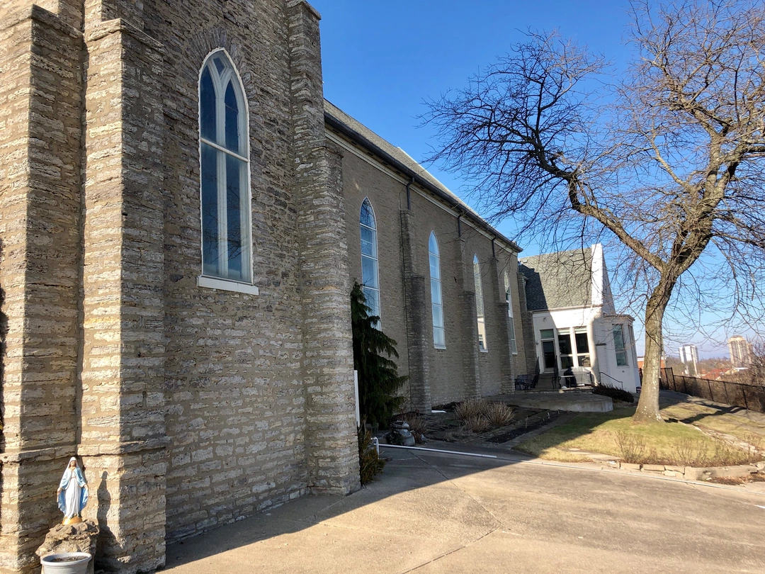 Gray stone Gothic-style church with tall arched windows and a statue of the Virgin Mary at the corner, with a white annex and bare trees under a blue sky.