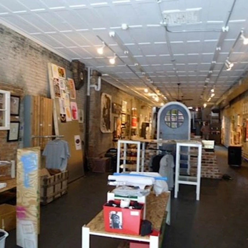 Long gallery room with exposed brick walls, a white tiled ceiling with track lights, and tables and a counter holding prints, boxes and clothing.