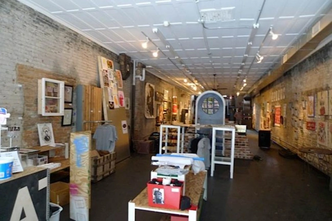 Long gallery room with exposed brick walls, a white tiled ceiling with track lights, and tables and a counter holding prints, boxes and clothing.