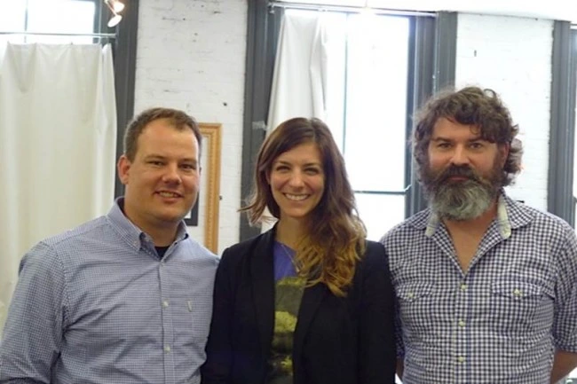 Three people standing side by side in a loft with white walls and curtains: a smiling man in a checked shirt, a smiling woman in a dark blazer, and a bearded man in a checked shirt.