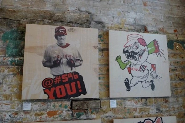 Two square wood-panel paintings on a brick wall: a baseball player in a Cincinnati Reds jersey above the words “@#$% YOU!”, and a cartoon mascot figure in a Reds uniform.
