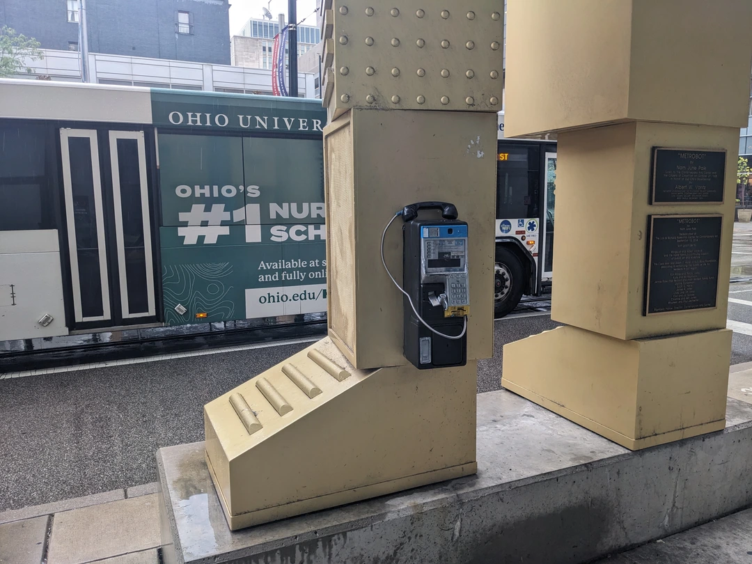 Beige steel base of a sculpture with a pay phone mounted on its front and two plaques on the columns, on a city sidewalk.